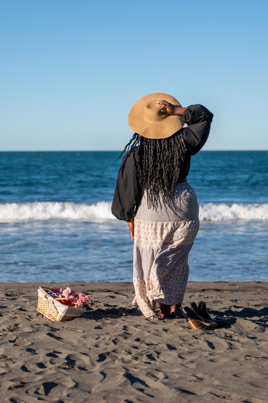 Khya owner, Kadijatu, on a beach wearing a wide-brimmed hat and long dress, with a basket of flowers on the sand.