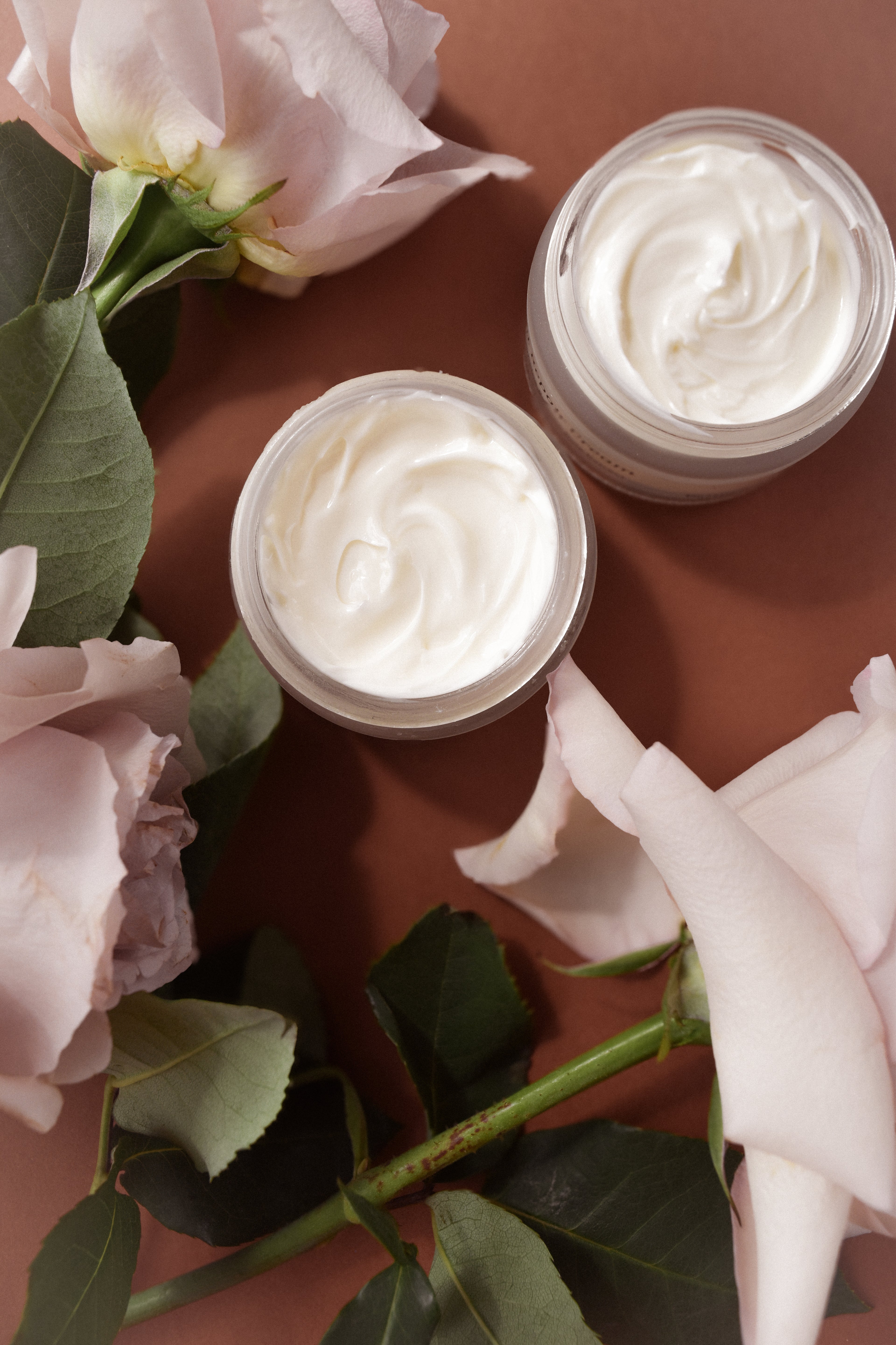Two jars of cream surrounded by pink flowers on a brown background